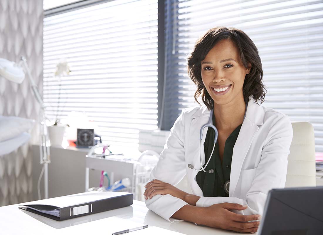 Medical Practice Insurance - Portrait of Smiling Female Doctor Wearing White Coat with Stethoscope Sitting behind Her Desk in the Office