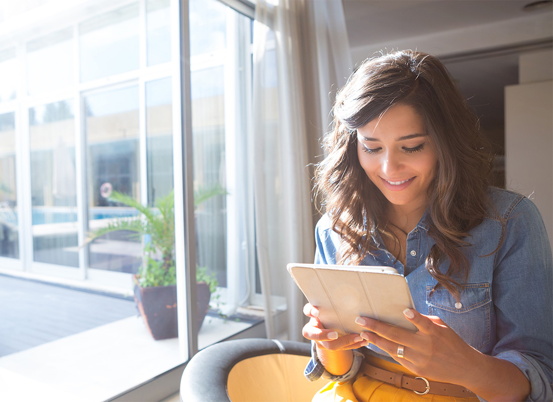 Blog - Portrait of a Young Smiling Woman Sitting Next to a Bright Window in the Office While Reading on a Tablet